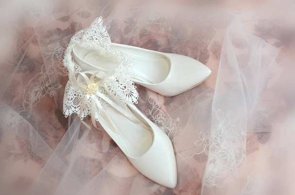 White wedding shoes and the bride's garter under a delicate veil on a blurry background - Fotografie, Obrázek