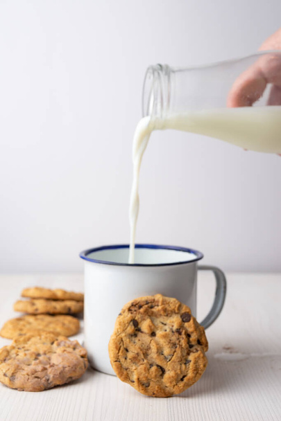Close-up of woman's hand with bottle, pouring milk over white cup and chocolate chip cookies, on white background, vertical - Foto, imagen
