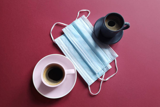 Surgical ear mask and a cup of coffee on a burgundy background. Medical mask protects against the virus of the crown or quarantine COVID-19. Biological concept of life. Lifestyle and Flatlay. - 写真・画像