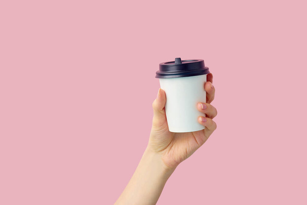 Female hand holding empty white blank paper cup of coffee with cap on pink background  - Foto, Imagen