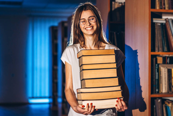 Young girl student with glasses in library smiling and carries stack of books. Exam preparation. - Фото, зображення