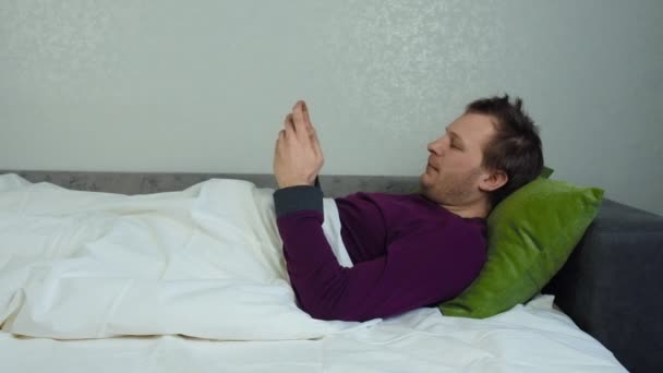 Man covered with a blanket is lying on the bed and playing a mobile game. Insomnia concept, sleeplessness. Caucasian unshaven guy. Mid shot slider movement, tracking left - Imágenes, Vídeo