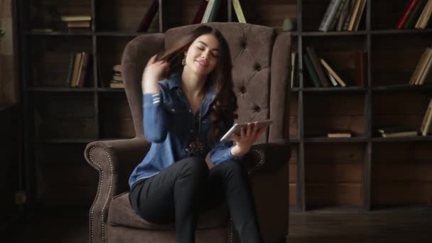 Young attractive woman sitting on a posh armchair with the tablet in her hands in the library - Materiał filmowy, wideo