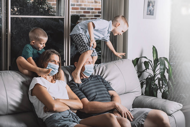 Mom and dad are wearing facial surgical masks. Two little sons are jumping over parents sitting on the couch trying to watch TV. - Photo, Image