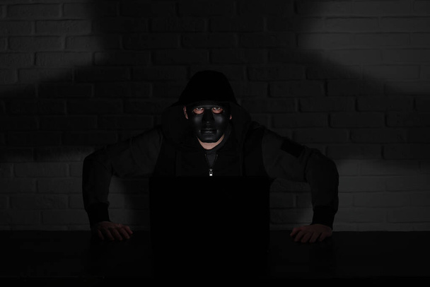 Hacker em máscara preta e capuz na mesa em frente ao monito - Foto, Imagem