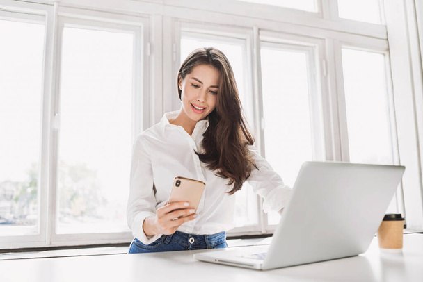 Woman using laptop and smartphone in office. Beautiful girl texting on phone. Entrepreneur, businesswoman, freelance worker, student working on computer. Business, technology concept - Фото, зображення