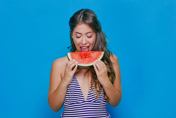 Pretty happy girl in a striped t-shirt eating watermelon on a blue background - Photo, Image