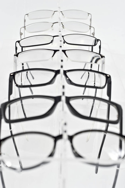 Many glasses on a white background. Glasses are stacked in a row on a transparent shop window. Glass stand on gray table. Sale and exhibition of optics. - Foto, imagen