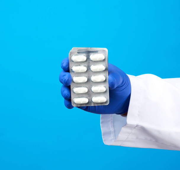 hand with blue sterile gloves holds a pills in a blister, concept for drug treatment of diseases, blue background - Photo, image