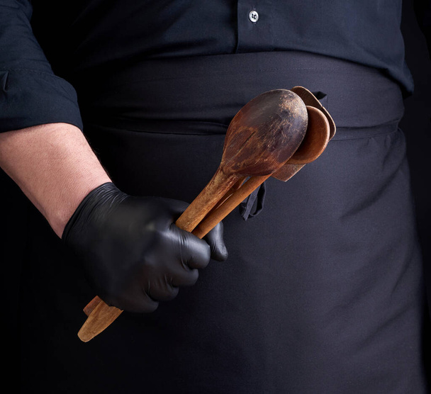 Koch in schwarzen Latex-Handschuhen und schwarzer Uniform hält hölzerne Vintage-Löffel vor sich, zurückhaltend - Foto, Bild