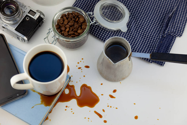 Coffee spilled on a book on the desktop. A cezve for coffee, coffee beans, a white cup of coffee, spill a drink on the table, making a puddle and ruining the book. Kitchen table, cook. - Foto, Imagem