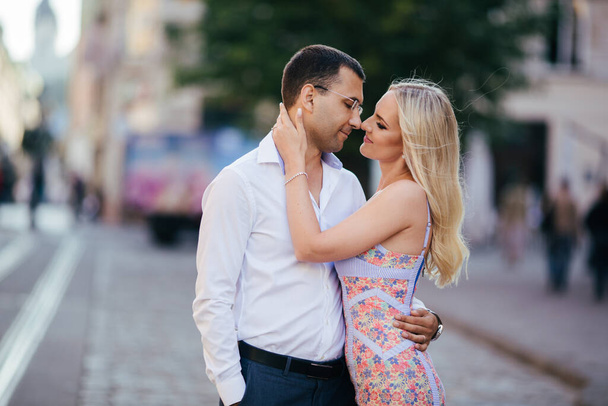 Lovely couple walking around the block. Dark-haired man in a white shirt hugging a blonde in a beautiful dress - Foto, imagen