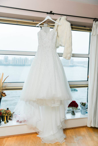 A vertical shot of a white wedding dress hanging from the wall with the window on the background - 写真・画像
