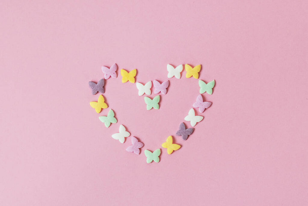 La masa multicolor sprinkles-mariposas se presentan en la forma de un corazón sobre un fondo rosa. Decoración de productos de confitería
 - Foto, imagen