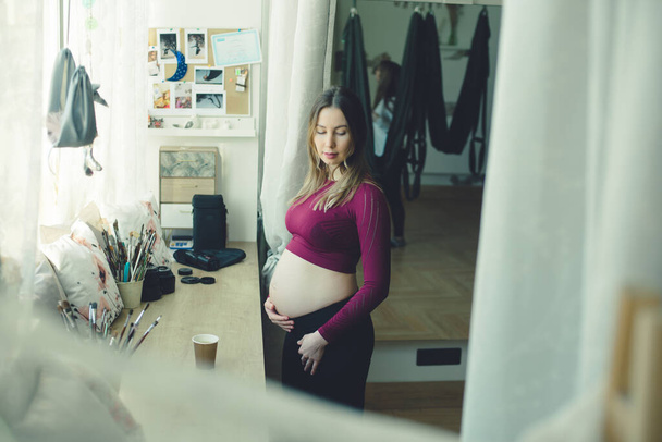 A pregnant woman drinks tea by the window. She wears tight sport costume, casual style. She hugs her baley and is very  carefull with future baby. Concept of mother care, young mother, art, drawing future, happy woman and child, healthy life. - Фото, изображение