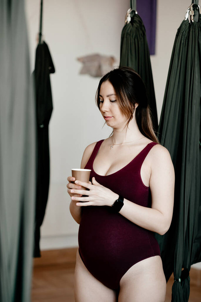 A pregnant woman is doing yoga in studio. She wears tight body. She hugs her baley and is very  carefull with future baby. Concept of mother care, young mother, woman and child, healthy life, fitness.  - Fotografie, Obrázek