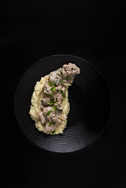  Lunch beef stroganoff met aardappelpuree en groene uien op zwarte plaat en op zwarte achtergrond - Foto, afbeelding