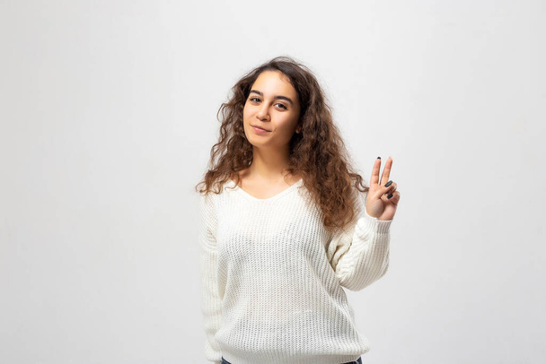 Young woman showing victory sign isolated on white background or woman showing peace sign. Beautiful young woman portrait. Indoor portrait of beautiful brunette young woman with shaggy hairstyle. - Photo, image
