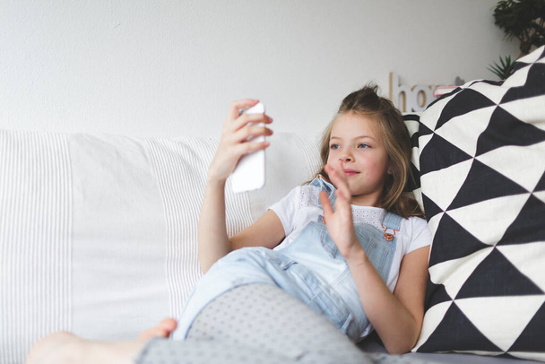 young girl with smartphone on couch is doing video call during the corona crisis - 写真・画像