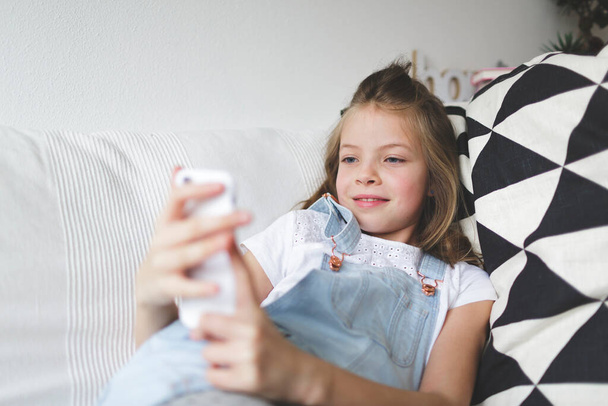 young girl with smartphone on couch is doing video call during the corona crisis - 写真・画像