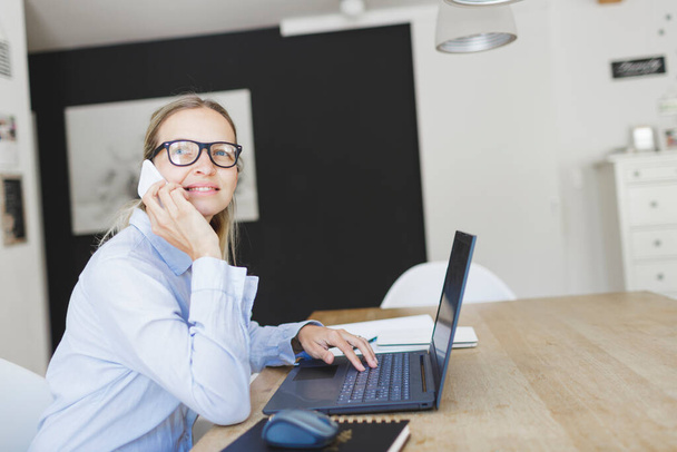 pretty young businesswoman with blue shirt and glasses working in home office and talking by phone - Valokuva, kuva