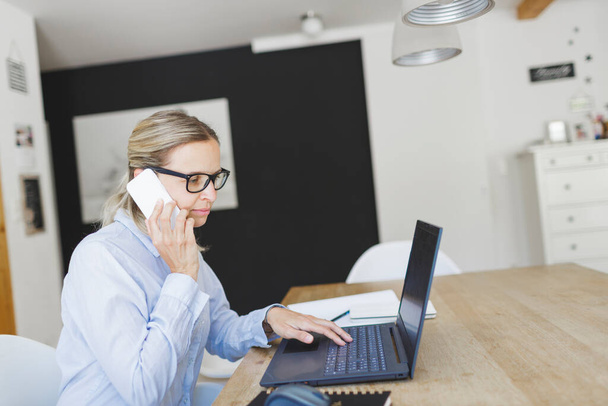 mujer de negocios bastante joven con camisa azul y gafas trabajando en la oficina en casa y hablando por teléfono
 - Foto, Imagen