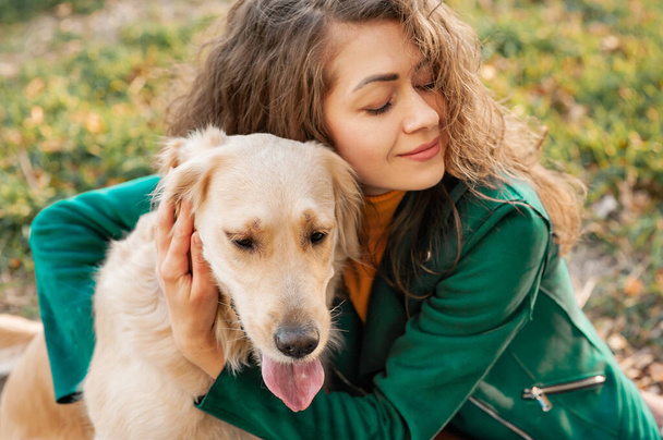 Riccia donna caucasica Ritratto che abbraccia il suo cane golden retriever nel parco estivo. Sfondo colore verde. Giovane donna riccia seduta con il suo cane all'aperto - Foto, immagini