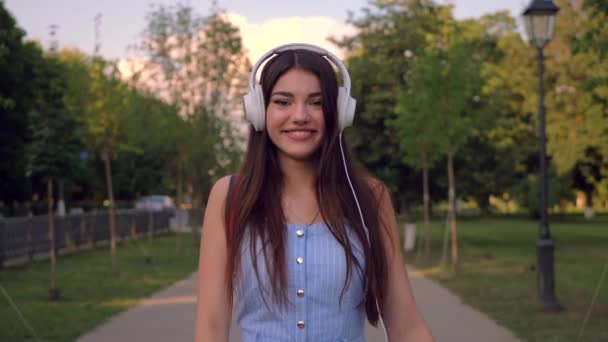 Enchanting lady is walking outdoors in park in summertime, listening to music in her headphones. Stylish woman is wearing blue romper suite.  - Filmati, video
