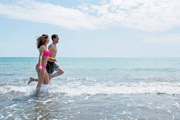 νεαρό ζευγάρι που διασκεδάζουν μαζί ενώ τρέχουν και πηδούν στο νερό στην παραλία  - Φωτογραφία, εικόνα