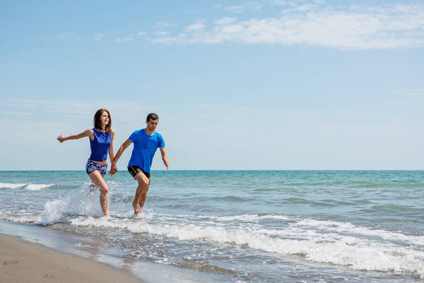 νεαρό ζευγάρι που διασκεδάζουν μαζί ενώ τρέχουν και πηδούν στο νερό στην παραλία  - Φωτογραφία, εικόνα