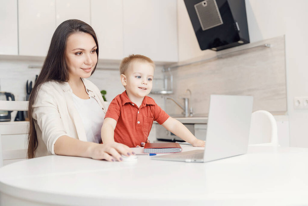 Frau im Mutterschaftsurlaub arbeitet online mit Laptop in der Küche mit kleinem Kind. Konzeptarbeit während der Quarantäne während Covid-19 - Foto, Bild