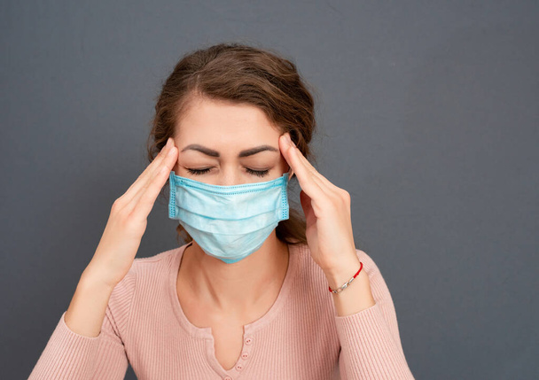 young brunette girl in medical mask holds her head on gray background. Female have pain because of severe headache, migraine. Concept face emotion - Photo, image