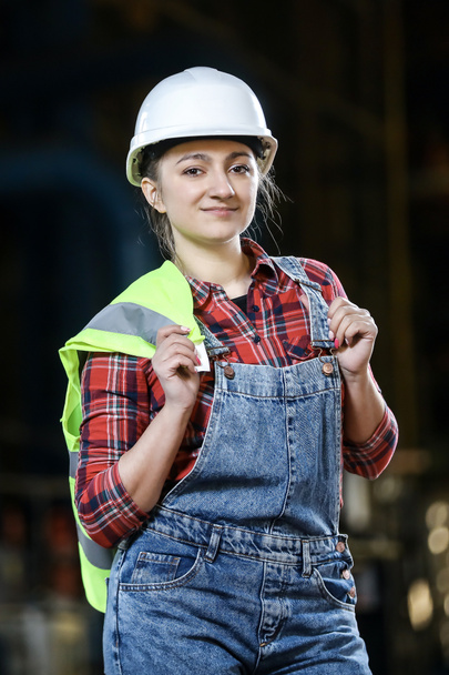 Mladá dívka v pracovních šatech a bílý pevný klobouk v továrně. Žena v pracovní uniformě, držící reflexní vestu ve skladu. Pracovní proces.  - Fotografie, Obrázek