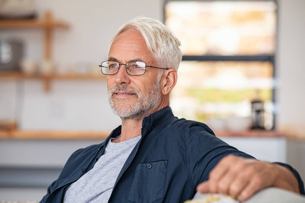 Retired mature man sitting on couch wearing spectacles and thinking. Smiling and cheerful old man planning the retirement. Thoughtful senior business man relaxing at home while looking away with copy space.  - 写真・画像