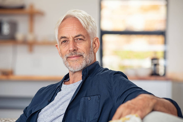 Handsome senior man with white hair sitting on couch and looking at camera. Casual satisfied old man relaxing on sofa while looking at camera. Portrait of proud mature man stay at home with copy space. - Фото, зображення