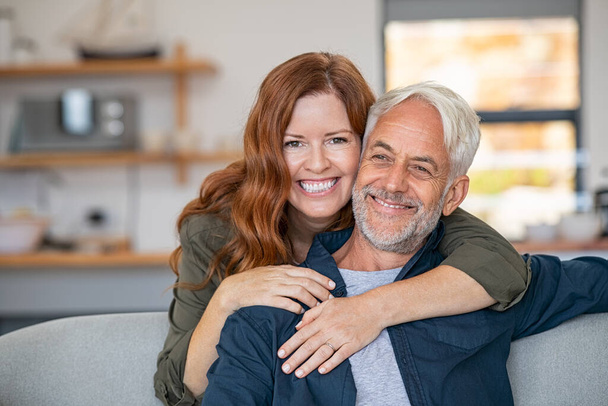 Smiling woman hugging her senior husband on couch from behind in the living room. Loving retired couple looking at camera and smiling at home. Portrait of smiling woman embracing mature man on sofa. - Photo, image