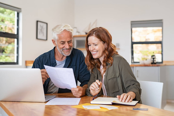 Mature smiling couple sitting and managing expenses at home. Happy senior man and mid woman paying bills and managing budget. Middle aged couple checking accountancy and bills while looking at receipt. - Valokuva, kuva