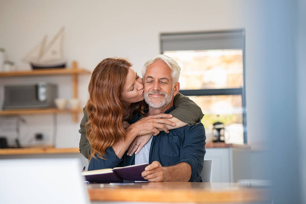 Happy loving woman kissing old man on cheek at home. Beautiful mature wife kissing her husband with beard and closed eyes. Senior couple in love. - Foto, afbeelding