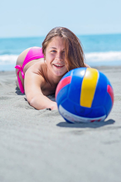 Genç bir kadın güneşli bir günde plajda pembe mayo içinde voleybol topuyla yatıyor. - Fotoğraf, Görsel