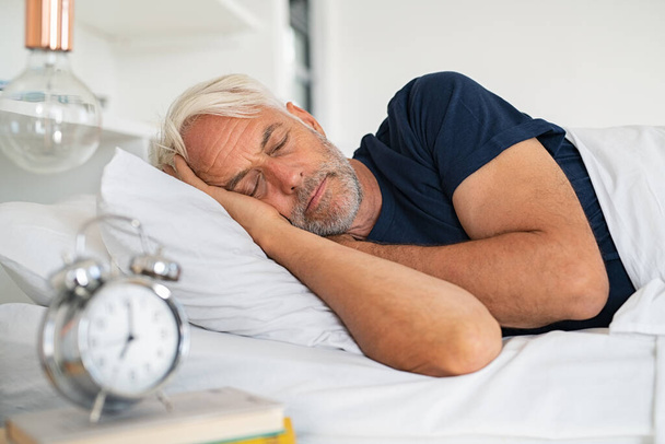 Old tired man sleeping on bed at home. Senior man with white hair in deep sleep on soft pillow at home. Mature healthy guy lying on side resting at home while sleeping. - Zdjęcie, obraz