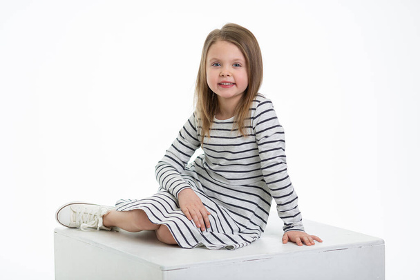 A small little blond girl in simple clothes with a cute smile sits full-length and looks at the camera Isolated on a white background. Beautiful smile of a child. Portrait of a happy laughing girl. - Фото, зображення