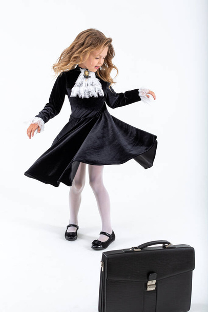 Cute young blonde schoolgirl in a beautiful school velvet black dress is spinning on an isolated white background. School time. Little beautiful girl in school uniform with blond hair smiling happy. - Foto, Bild
