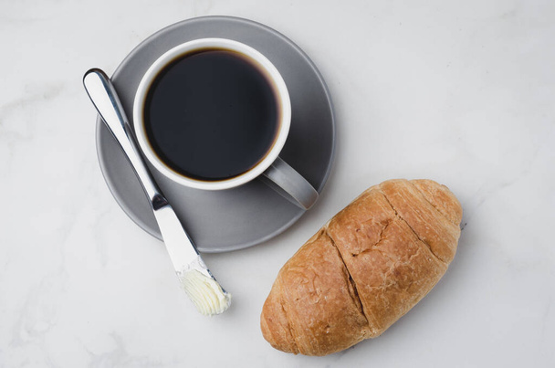 Pausa para el café con cuchillo de mantequilla de croissant y taza de café negro en una mesa de piedra blanca. Vista superior
. - Foto, imagen