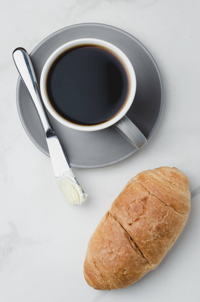 Coffee break with croissant butter knife and cup of black coffee on a white stone table. Top view. - Фото, зображення