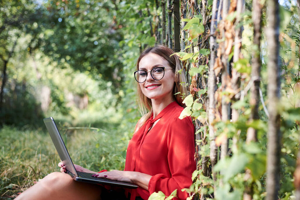Young pretty blond woman, wearing red shirt and eyeglasses, sitting in the garden holding laptop on her lap. Student, working studying with notebook in the park in summer. Campus leisure time. - Фото, изображение
