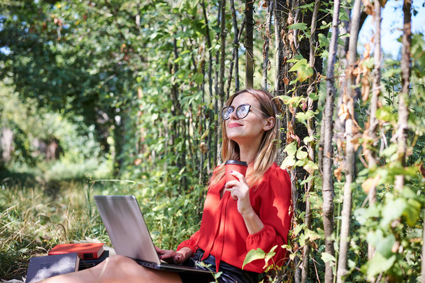 Young pretty blond woman, wearing red shirt and eyeglasses, sitting in the garden holding laptop on her lap. Student, working studying with notebook in the park in summer. Campus leisure time. - Foto, afbeelding