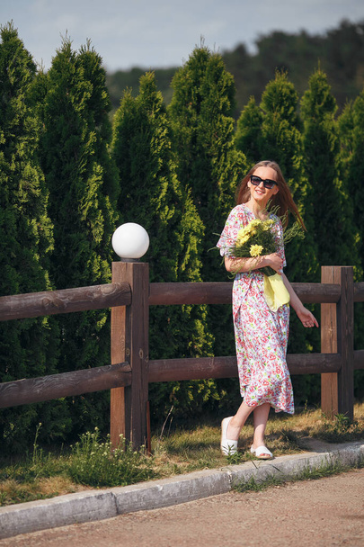 Fille élégante posant dans une robe dans un parc printanier ensoleillé. Portrait calme d'une belle fille debout avec un bouquet au printemps - Photo, image