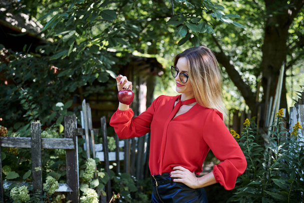 Young blond woman, wearing eyeglasses and red blouse, holding red claret apple in her hand. Three-quarter picture of female student in the garden in spring. An apple a day keeps doctor away. - Valokuva, kuva