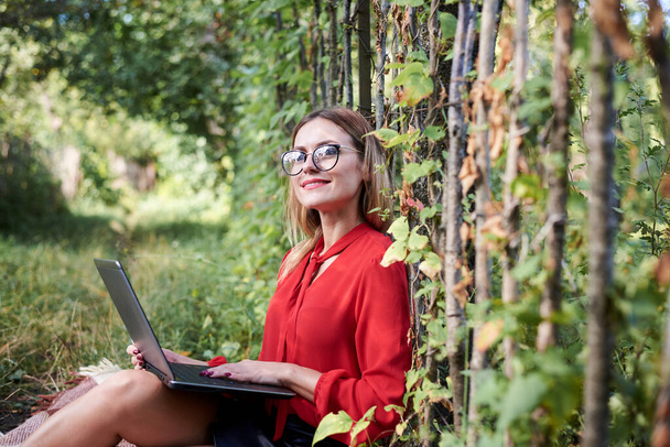 Young pretty blond woman, wearing red shirt and eyeglasses, sitting in the garden holding laptop on her lap. Student, working studying with notebook in the park in summer. Campus leisure time. - Fotó, kép