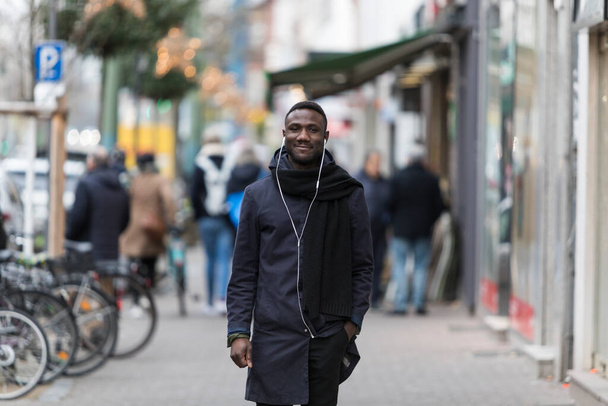 Junger schwarzer Mann läuft glücklich durch die Stadt und lächelt mit einer Hand in der Tasche. - Foto, Bild